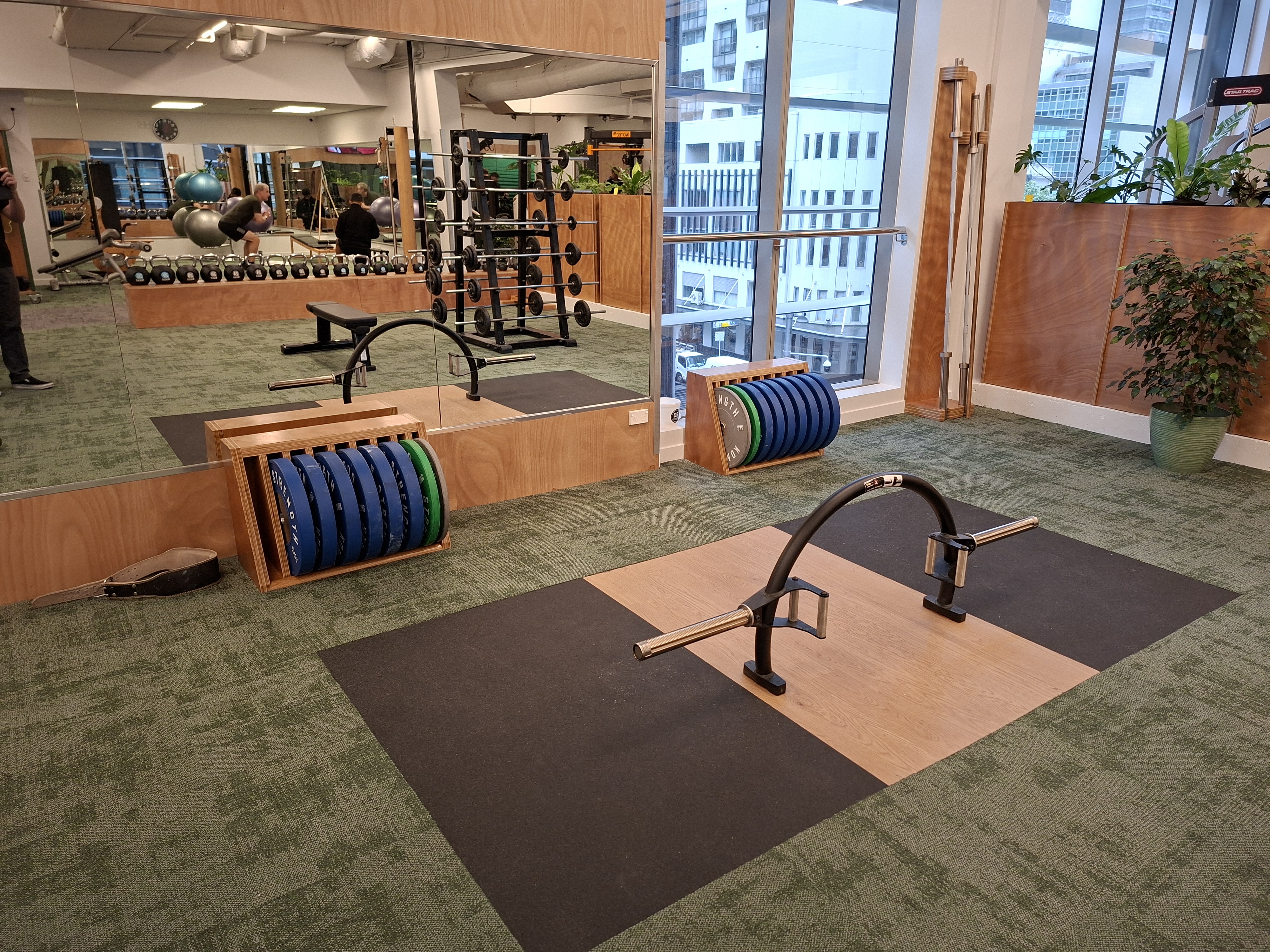 Hip hinge zone deadlift platform with blue bumper plates and trap bar, mirror reflecting kettlebells and wider training floor